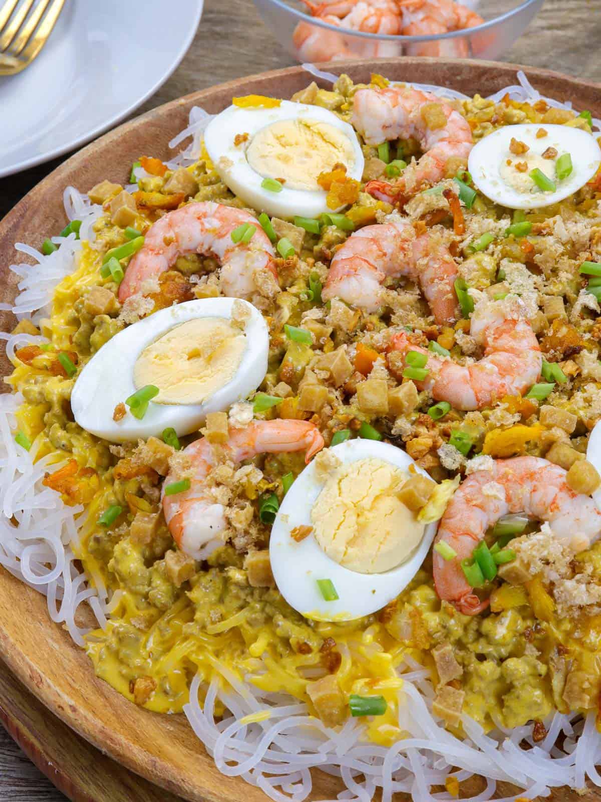 Creamy Palabok on a wooden platter with shrimp, hardboiled eggs, green onions, fried tofu toppings.