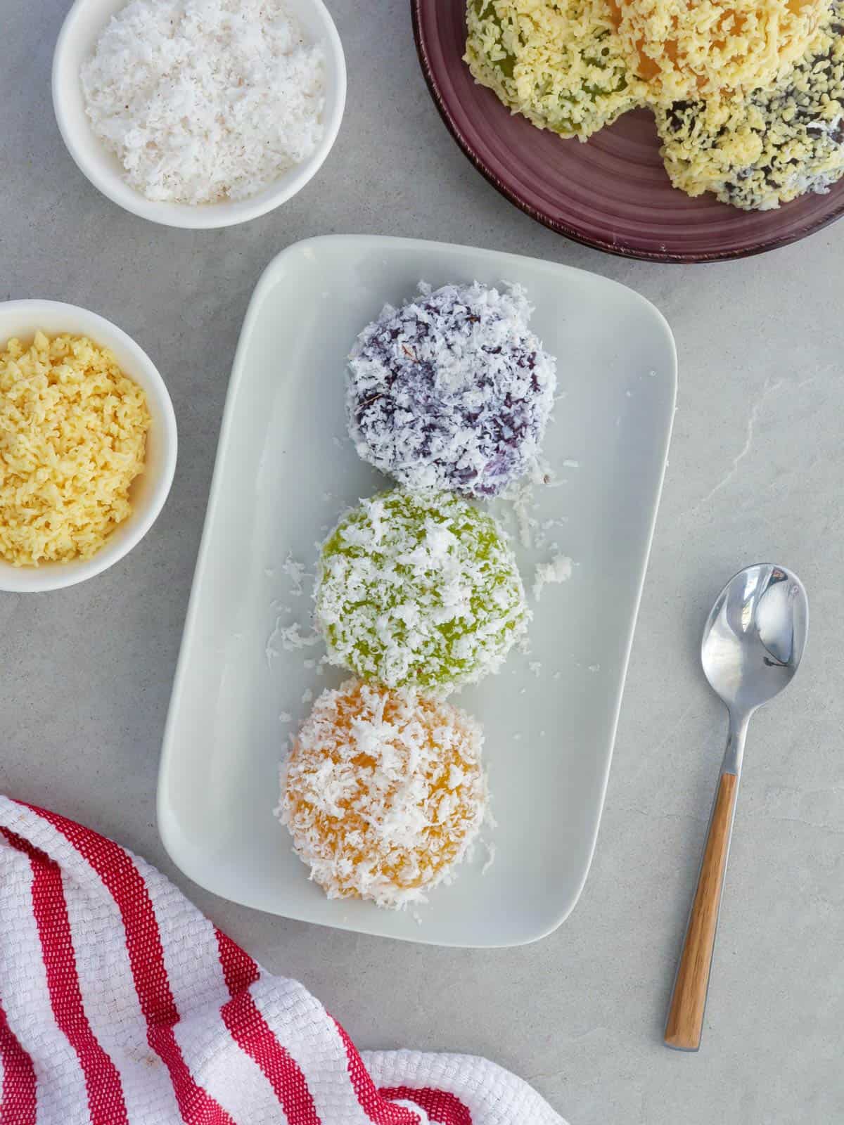 pichi-pichi with different flavors and coated with grated coconut on a white platter with a bowl of shredded cheese in the background.
