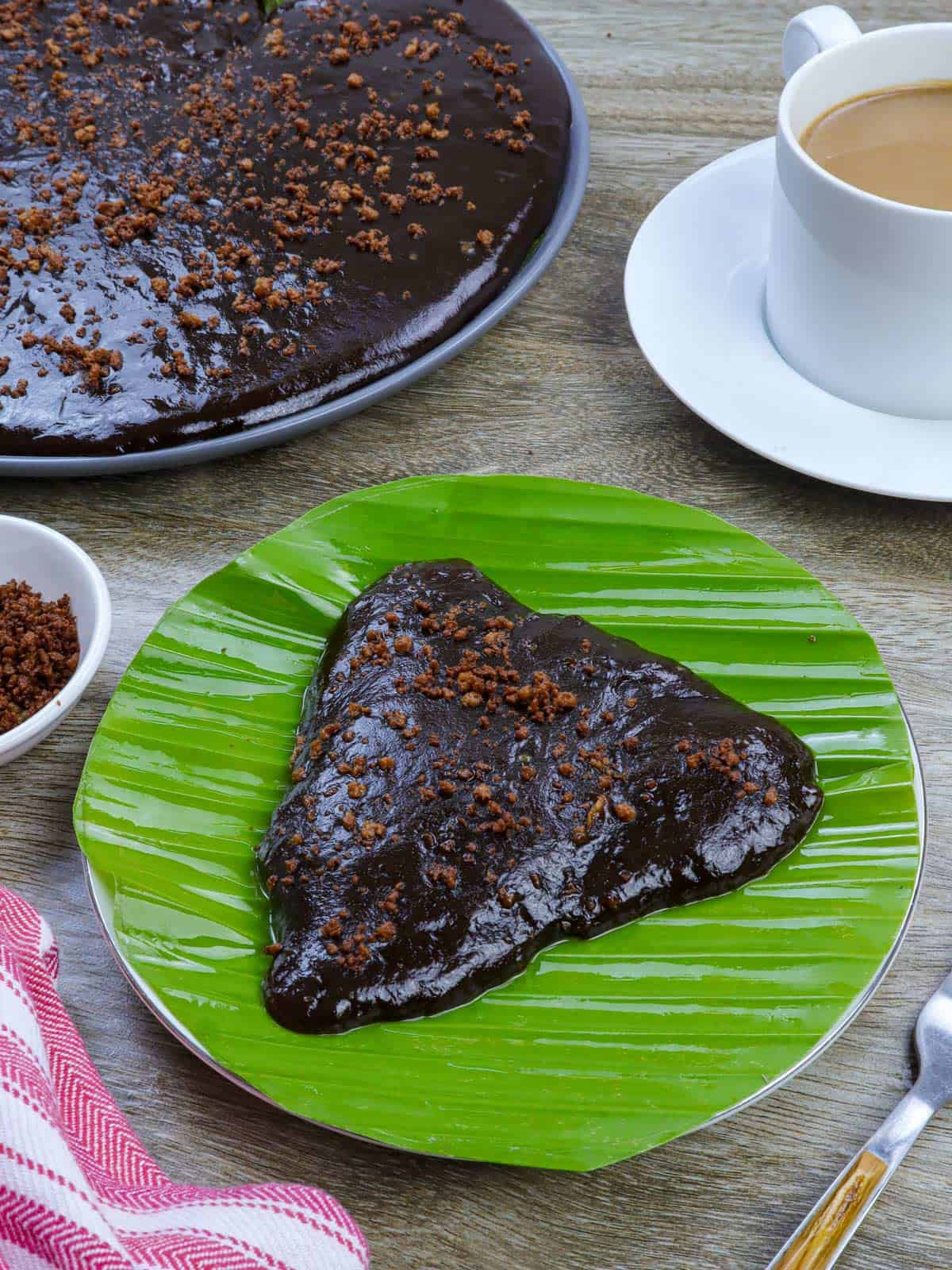 slice of Kalamay Hati on a banana leaf lined plate with a cup of coffee in the background.