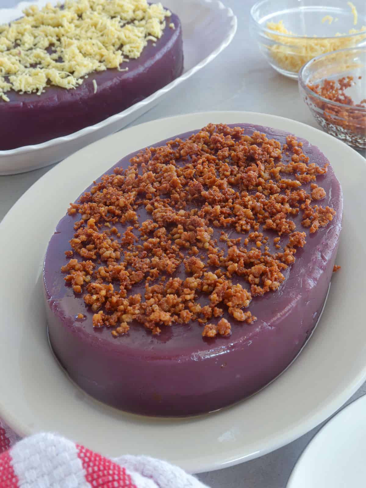 ube halaya topped with latik on a white serving platter with a ube halaya topped with cheese in the background.